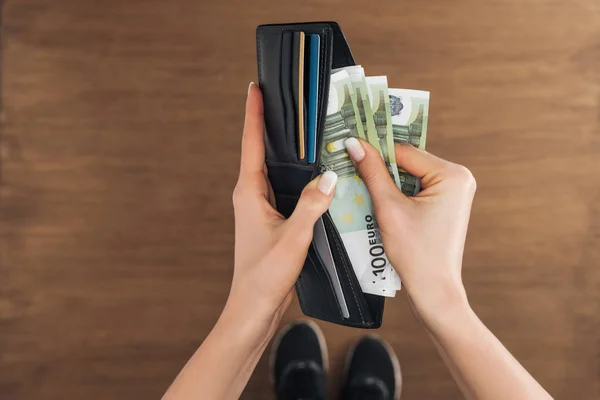 Vista superior de la mujer poniendo billetes en euros en la cartera sobre fondo de madera - foto de stock