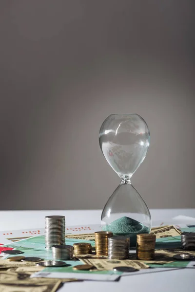 Reloj de arena y pilas de monedas en diferentes billetes con fondo gris - foto de stock