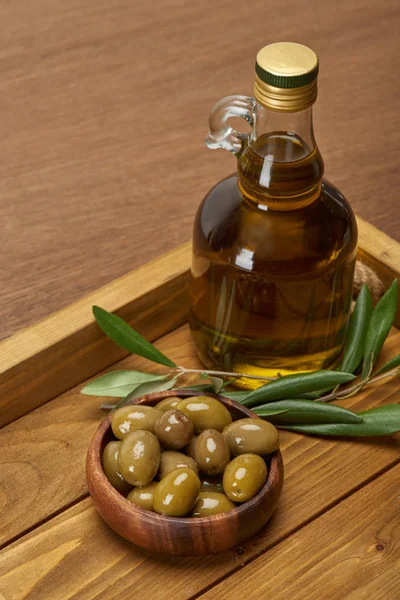 Bandeja de madera con botella de aceite, tazón de aceitunas y hojas de olivo en la superficie marrón - foto de stock