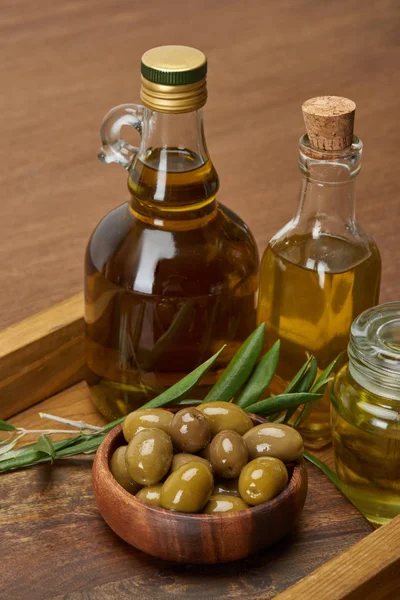 Wooden tray with different oil bottles, bowl of olives and olive tree leaves on brown surface — Stock Photo