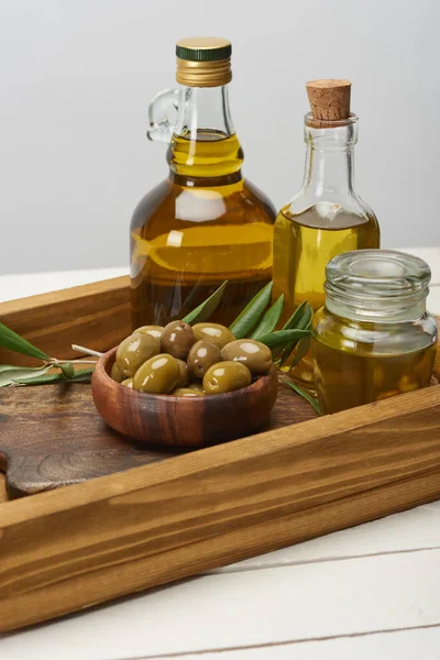Bandeja de madera con cuenco de aceitunas, botellas de aceite y hojas de olivo en la superficie blanca - foto de stock