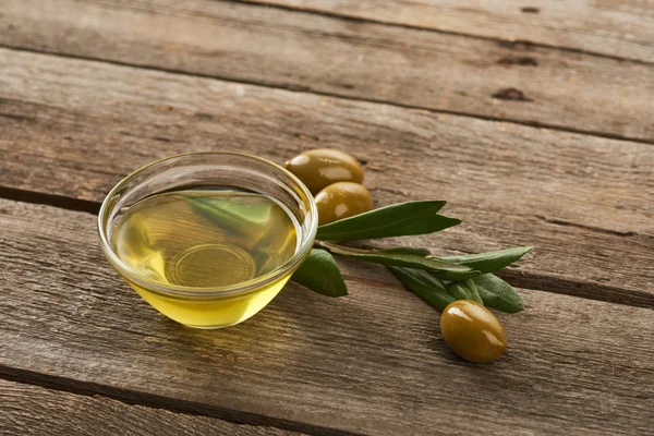Glass bowl with oil, olive tree leaves and olives on brown wooden surface — Stock Photo