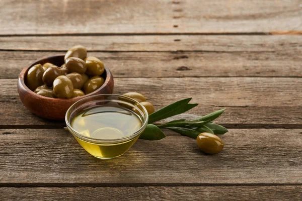 Tazón de vidrio de aceite, tazón de madera con aceitunas, hojas de olivo y aceitunas en la superficie de madera marrón - foto de stock