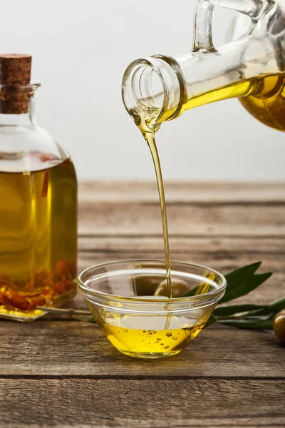Verser l'huile de la bouteille dans un bol en verre, une bouteille d'huile, des feuilles d'olivier et de l'olive sur une surface en bois — Photo de stock