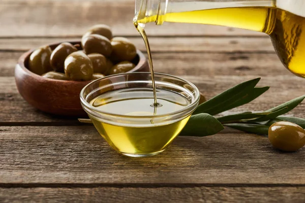 Verter el aceite de la botella en un recipiente de vidrio, un tazón de aceitunas, una rama de olivo y una aceituna sobre una superficie de madera - foto de stock