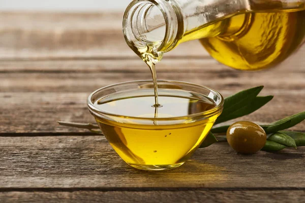 Pouring oil from bottle into glass bowl, olive tree leaves and olive on wooden surface — Stock Photo