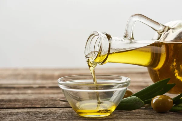 Öl aus der Flasche in Glasschale gießen, Olivenbaumblätter und Oliven auf Holzoberfläche — Stockfoto
