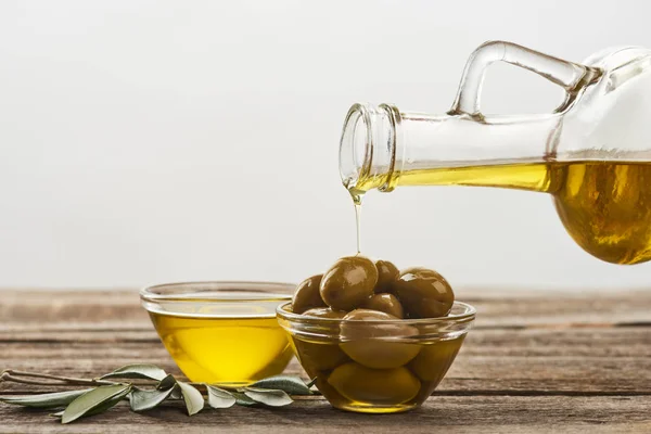 Verter en un tazón de vidrio con aceitunas, un tazón lleno de aceite y una rama de olivo sobre una superficie de madera - foto de stock