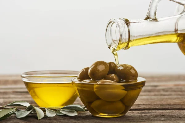 Verter aceite en un recipiente de vidrio con aceitunas, un recipiente lleno de aceitunas y una rama de olivo sobre una superficie de madera - foto de stock