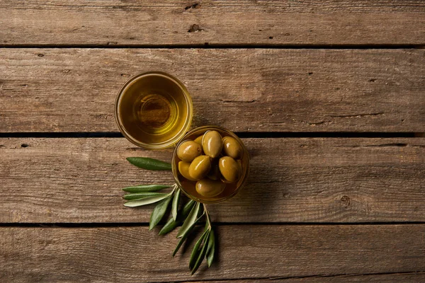 Vista superior de taças de vidro com azeitonas e óleo, e ramo de oliveira na superfície de madeira — Fotografia de Stock