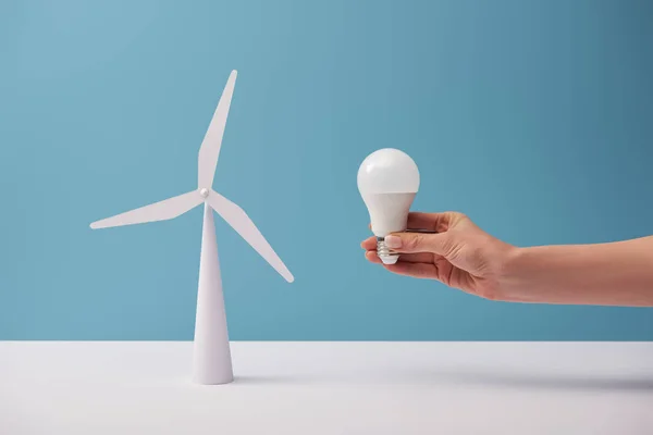 Vue recadrée d'une femme tenant une ampoule sur une table blanche avec un modèle de moulin à vent sur fond bleu — Photo de stock