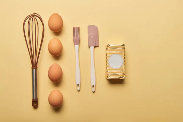 Puesta plana con batidor de globos, huevos, espátula, pincel de panadería y mantequilla sobre fondo amarillo - foto de stock
