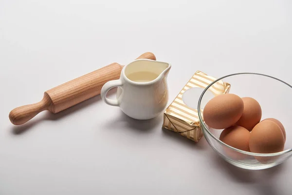 Rodillo, frasco con leche, mantequilla y huevos en un bol sobre fondo gris - foto de stock
