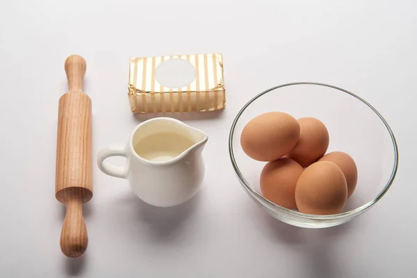 Rodillo, frasco con leche, mantequilla y huevos en un bol sobre una superficie gris - foto de stock