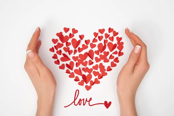 Vista recortada de las manos femeninas cerca de la disposición en forma de corazón de pequeños corazones cortados de papel rojo con letras de 