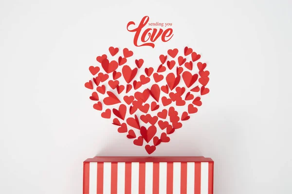 Top view of heart shaped arrangement of small red paper cut hearts and striped gift box on white background with 