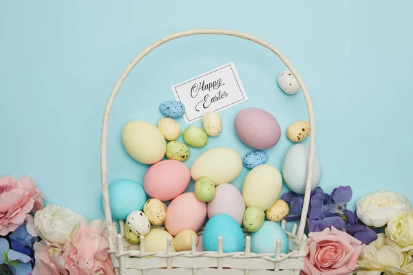 Vista superior de pollo pintado de Pascua y huevos de codorniz en canasta de paja, flores y tarjeta con letras de Pascua feliz - foto de stock