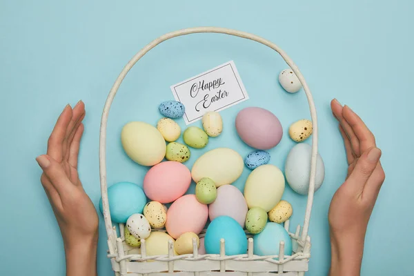 Vue recadrée de la femme tenant des œufs de poulet et de caille peints de Pâques dans un panier de paille, carte avec lettrage de Pâques heureux — Photo de stock