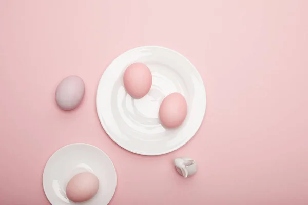 Top view of toy bunny, painted easter eggs on saucer and plate on pink background — Stock Photo