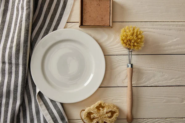 Вид сверху на пустую белую тарелку, полосатое полотенце и различные чистящие средства на белой деревянной поверхности, концепция нулевых отходов — стоковое фото