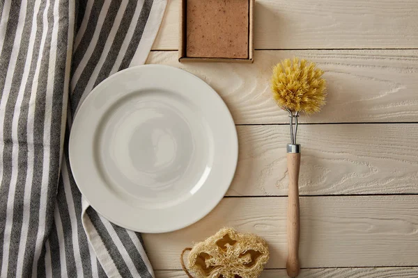 Top view of empty white plate, striped towel and different cleaning items on white wooden surface, zero waste concept — Stock Photo