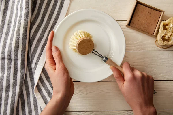 Partial view of woman washing dish, striped towel and cleaning items on white wooden surface, zero waste concept — Stock Photo