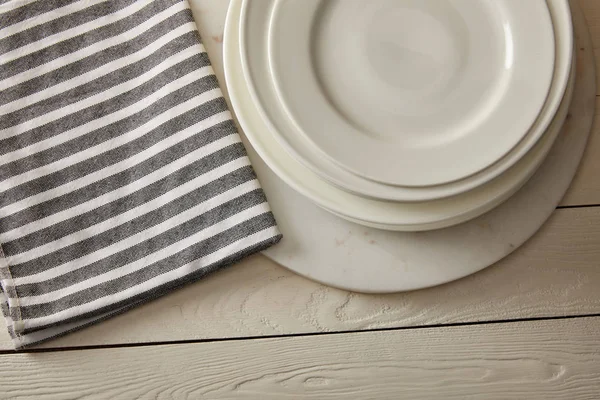 Vue de dessus des assiettes empilées et serviette en coton rayé sur la surface en bois blanc — Photo de stock