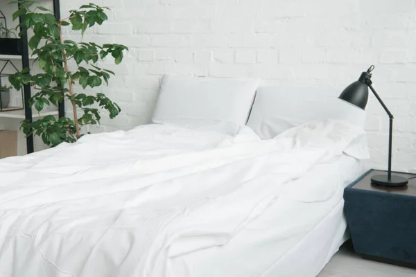 Cama con manta blanca y almohadas, planta y lámpara en la mesita de noche - foto de stock