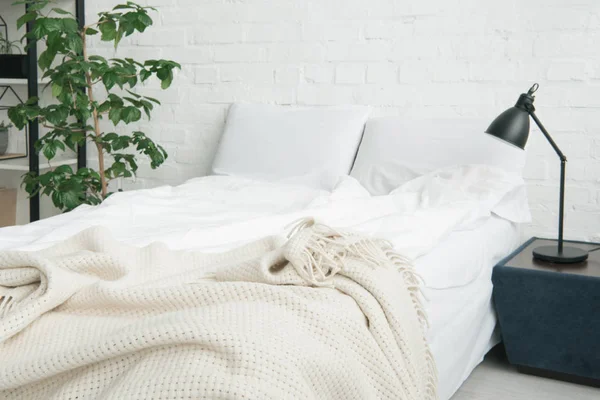 Cama com cobertor branco e travesseiros, planta e lâmpada na mesa de cabeceira preta — Fotografia de Stock