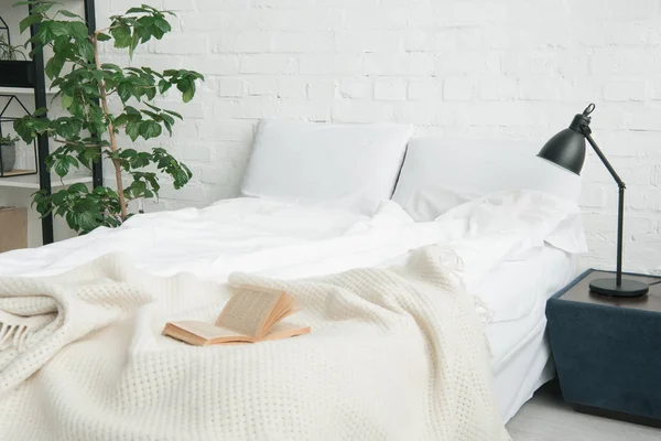 Libro sobre cama blanca, planta y lámpara sobre mesita de noche negra - foto de stock