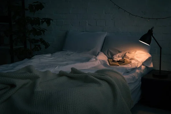 Интерьер спальни с книгой и стаканами на пустой кровати, растения и лампы на черном тумбочке ночью — стоковое фото