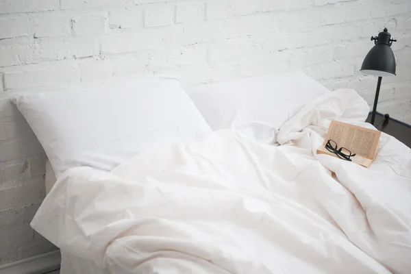 Brille und Buch auf dem Bett mit weißer Decke und Kissen, Lampe — Stockfoto