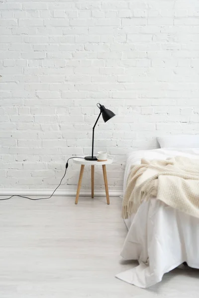 Intérieur de la chambre confortable avec oreiller sur lit, lampe et tasse de café sur tabouret — Photo de stock