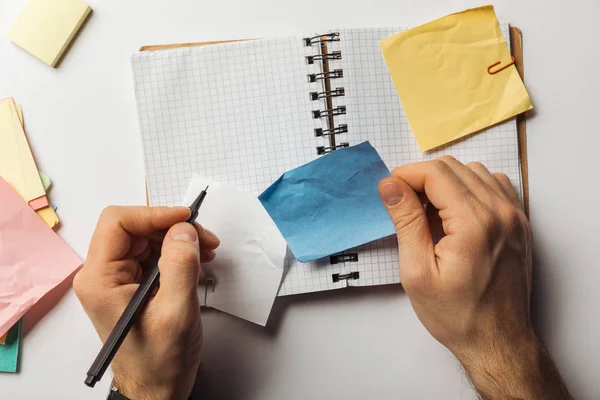 Cropped view of man writing on sticky note and holding crumpled blue paper near opened notebook — Stock Photo