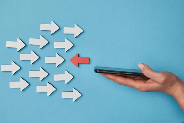 Cropped view of woman holding smartphone near red horizontal pointer opposite white arrows on blue background — Stock Photo