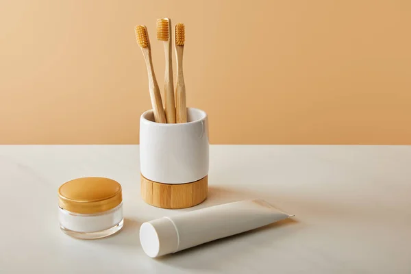 Spazzolini di bambù in supporto, dentifricio in tubo e crema cosmetica su tavolo bianco e fondo beige — Foto stock