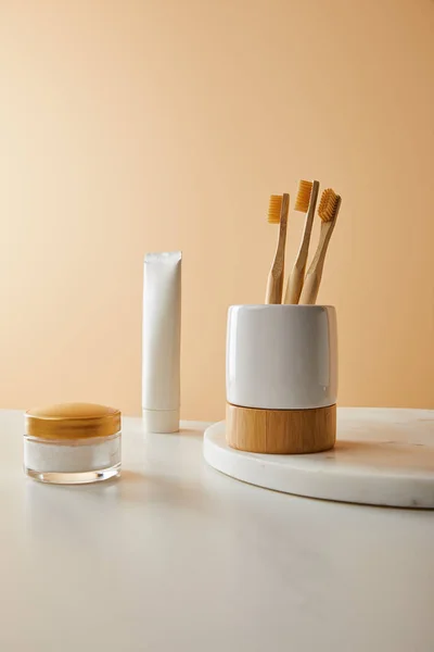 Soporte con cepillos de dientes sobre tabla redonda de mármol, pasta de dientes en tubo y crema cosmética sobre mesa blanca y fondo beige - foto de stock