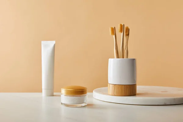 White holder with bamboo toothbrushes on marble round board, toothpaste in tube and cosmetic cream on table and beige background — Stock Photo