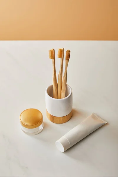Soporte con cepillos de dientes de bambú, pasta de dientes en tubo y crema cosmética sobre mesa de mármol blanco y fondo beige - foto de stock