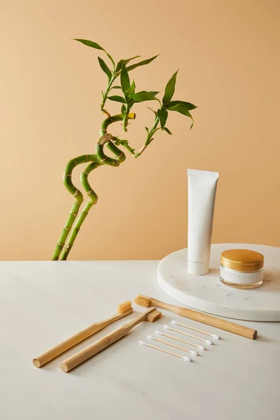 Toothpaste in tube, cosmetic cream, bamboo toothbrushes and ear sticks on table, green bamboo on beige background — Stock Photo