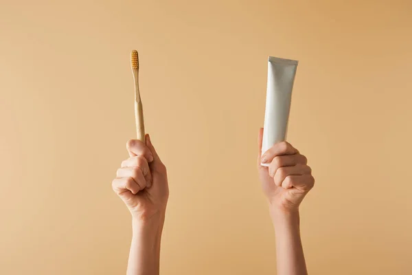 Vista recortada de la mujer sosteniendo el cepillo de dientes de bambú marrón y pasta de dientes en tubo sobre fondo beige - foto de stock