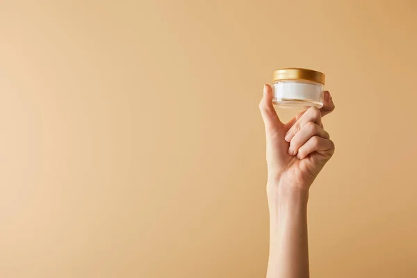Vista recortada de la mano femenina celebración crema cosmética sobre fondo beige - foto de stock