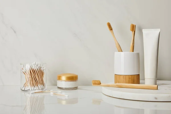 Pasta de dientes en tubo, soporte, cepillos de dientes de bambú, crema cosmética y orejeras en vidrio sobre fondo de mármol blanco - foto de stock