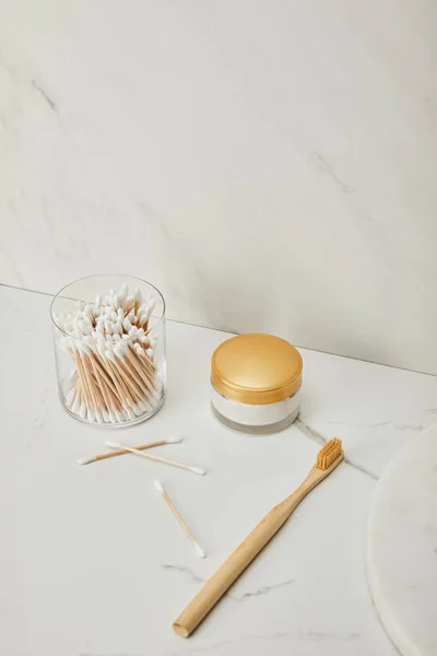 Varas de ouvido em vidro, creme cosmético e escova de dentes de bambu no fundo de mármore branco — Fotografia de Stock