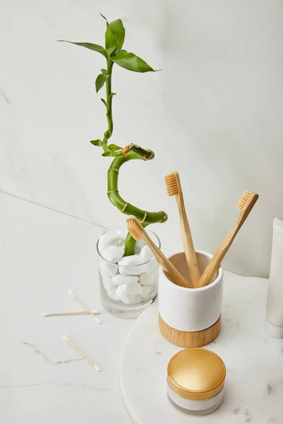 Planche ronde avec brosses à dents dans le support, crème cosmétique et dentifrice dans le tube près de tige de bambou vert dans un vase sur fond de marbre blanc — Photo de stock