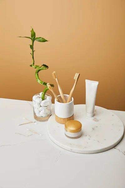 Escovas de bambu em suporte, creme cosmético e pasta de dentes em tubo em placa redonda perto de haste de bambu verde em vaso sobre fundo bege — Fotografia de Stock