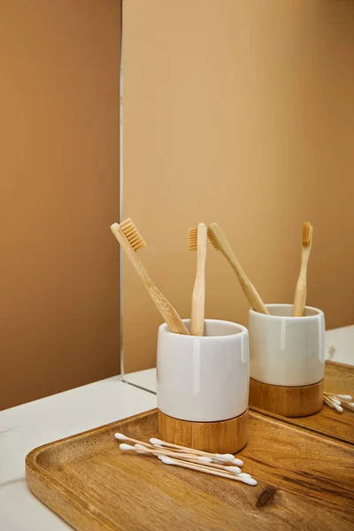 Tablero con cepillos de dientes de bambú en el soporte y los palos de la oreja al lado del espejo en la mesa blanca y fondo beige - foto de stock