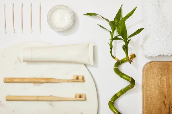 Vista superior da placa redonda de mármore com escovas de dentes de bambu e pasta de dentes em tubo, creme cosmético, auriculares, haste de bambu e toalha no fundo branco — Fotografia de Stock