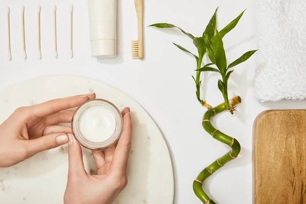 Abgeschnittene Ansicht einer Frau, die kosmetische Creme über ein Marmorrundbrett hält, Zahnbürste, Zahnpasta in der Tube, Ohrstöpsel, Bambusstamm und Handtuch auf weißem Hintergrund — Stockfoto