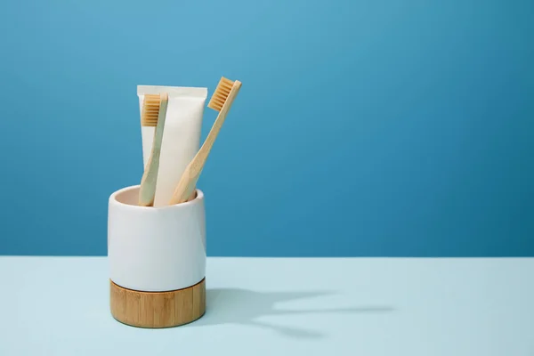 Soporte con pasta de dientes en tubo y cepillos de dientes de bambú sobre mesa y fondo azul - foto de stock